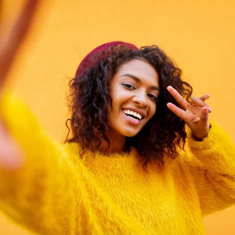 African woman with wavy hairs  making self portrait   over yellow urban wall.  Perfect  smile .  Stylish black girl shows signs by hand.